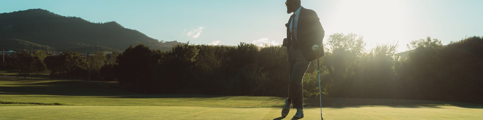 A silhouette of an elegant bald bearded black man in a tailored suit leaning on his gold club while standing on an empty lawn of a golf field, backlit by evening sun casting a long shadow on the grass