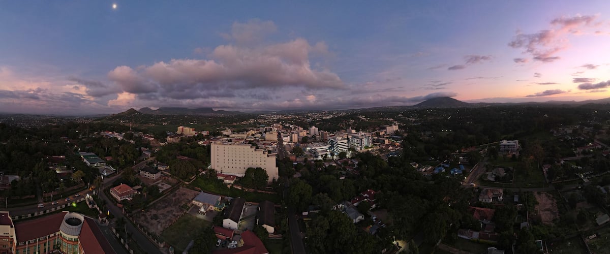 A cityscape at dusk with a prominent building in the center, surrounded by residential areas and a mountain in the background.