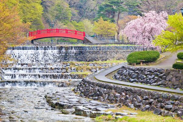 A red bridge over a cascading waterfall, surrounded by autumn foliage and a cherry blossom tree.