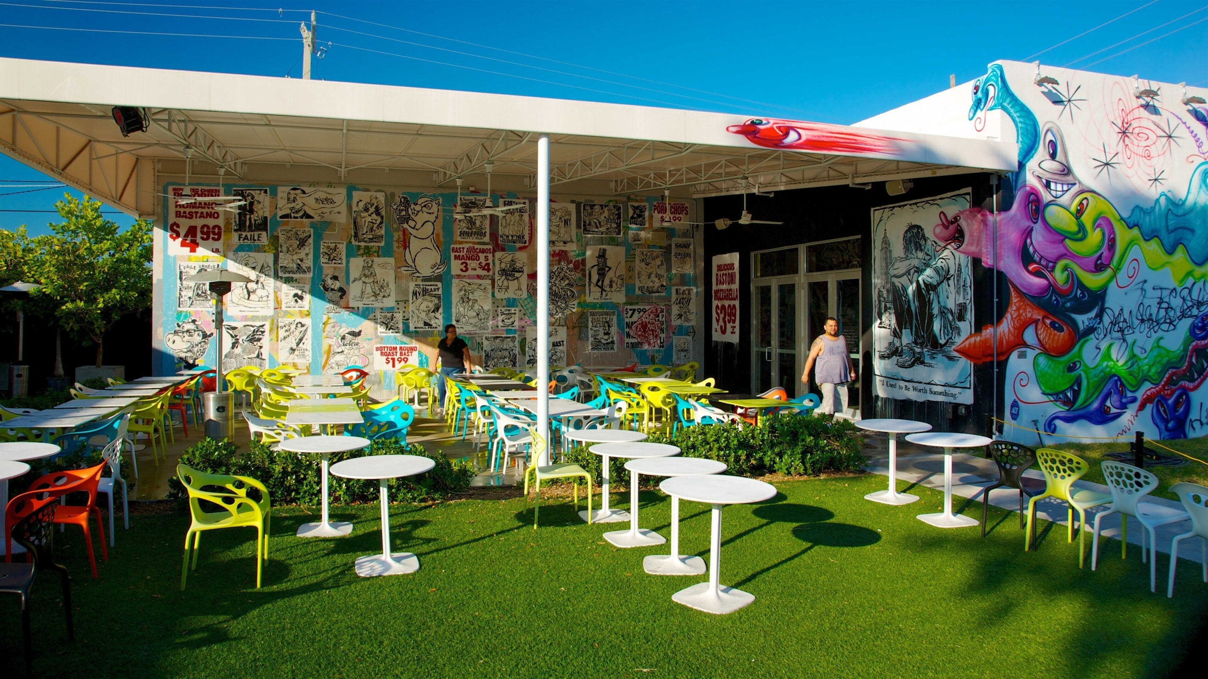 Uma área de refeições ao ar livre com cadeiras e mesas coloridas, cercada por um mural com diversas obras de arte e texto.