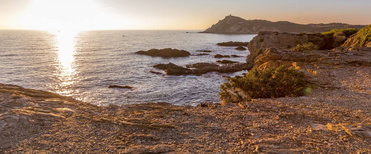 A rocky coastline with a lighthouse in the distance during sunset.