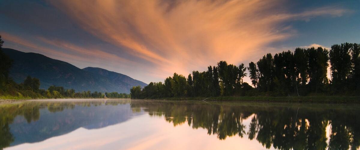 A calm lake reflecting trees and mountains under a colorful sky.