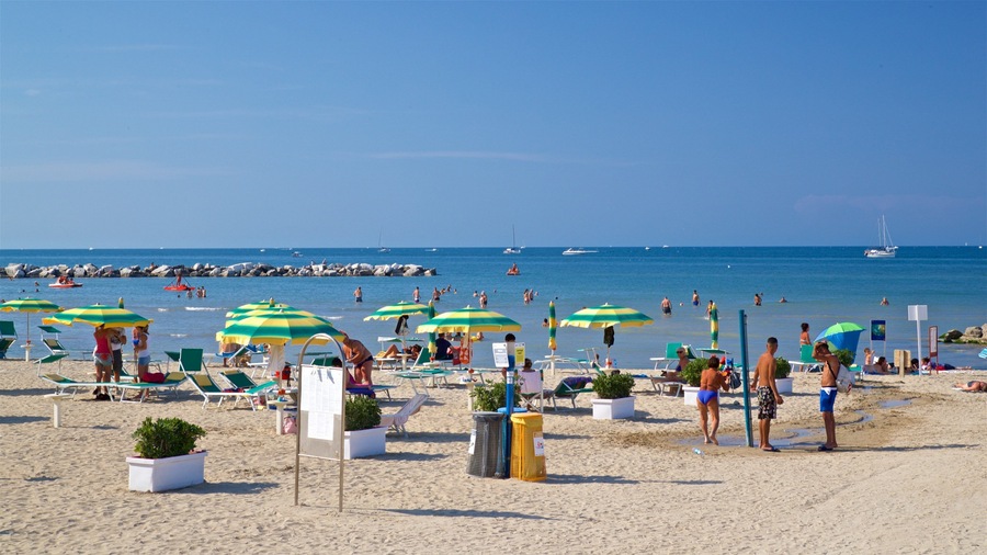 A sunny beach with people relaxing under umbrellas, a sandy shore, and boats in the water.