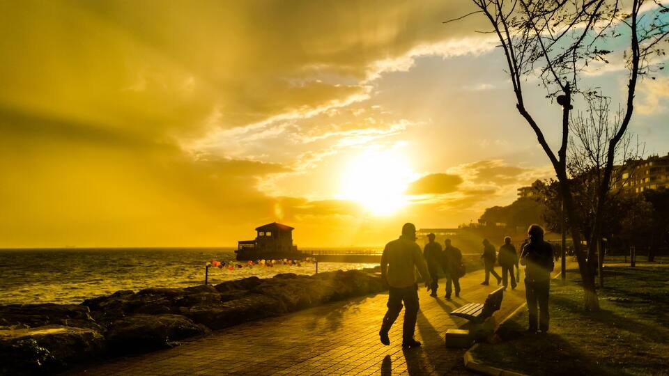 A sunset over a coastal area with a pier, people walking, and a building on the shore.
