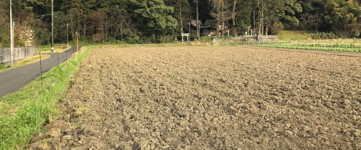 A rural landscape with a plowed field, a road, and a traditional building in the background.