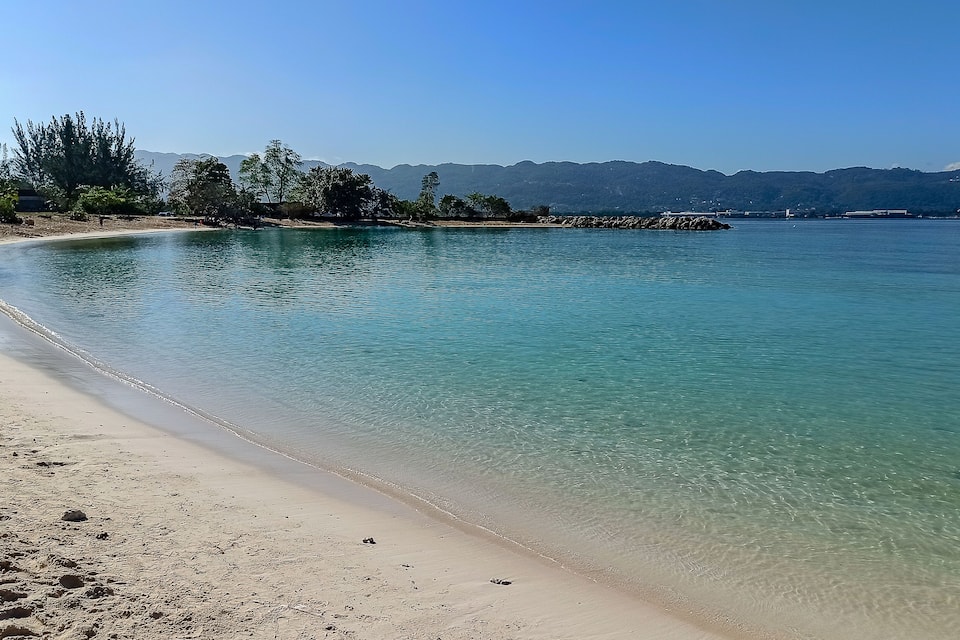 A clear beach with calm waters, sandy shore, and distant mountains.