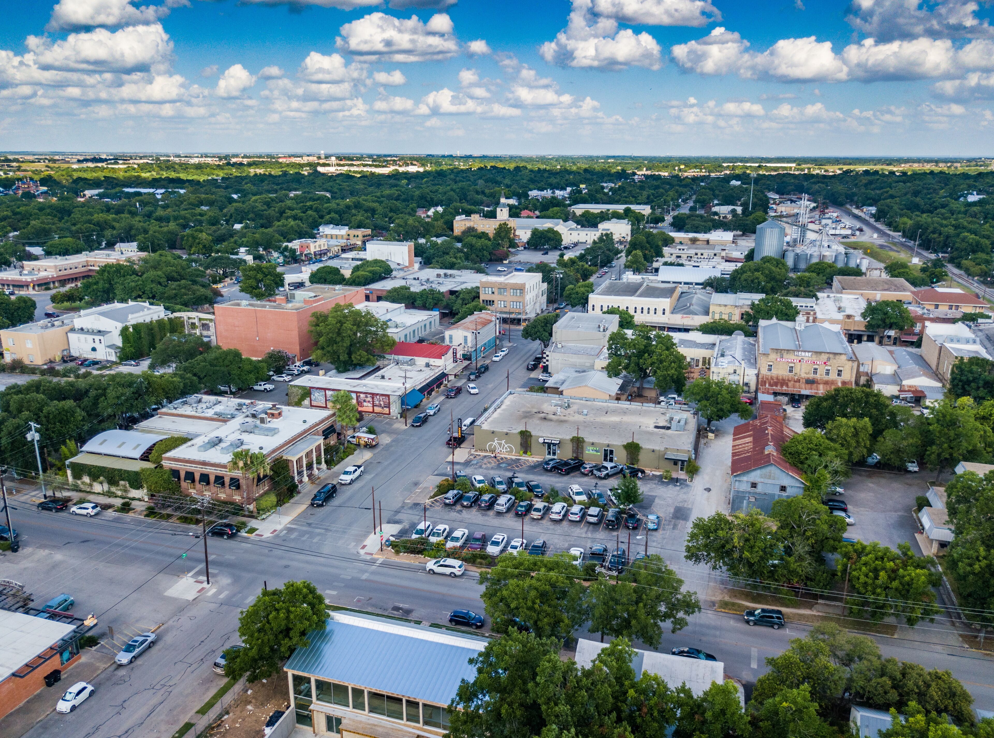 An aerial view of a town with a mix of historic and modern buildings, a central street, and surrounding greenery.