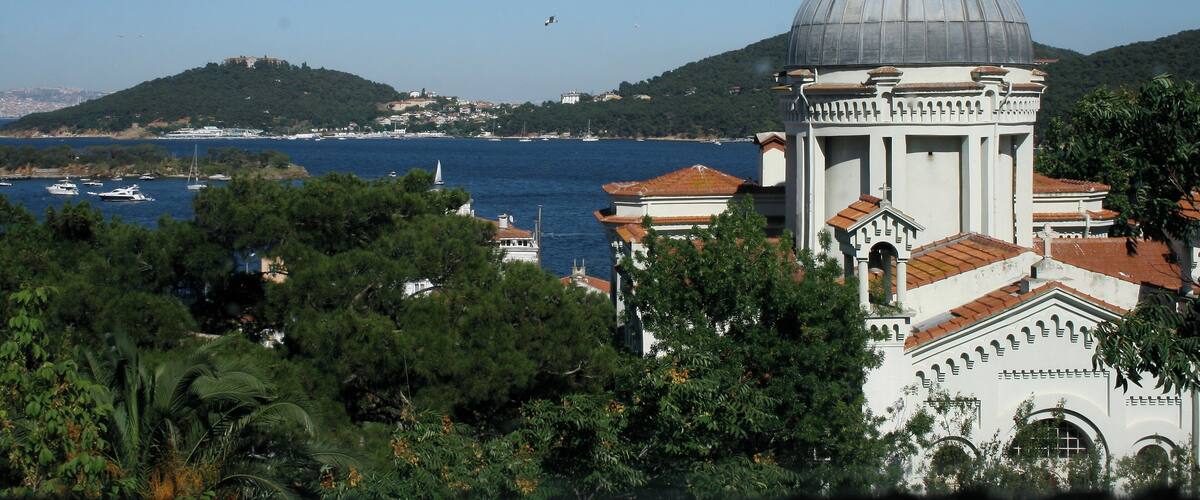 A coastal church with a dome, surrounded by lush greenery and a view of the sea with boats.