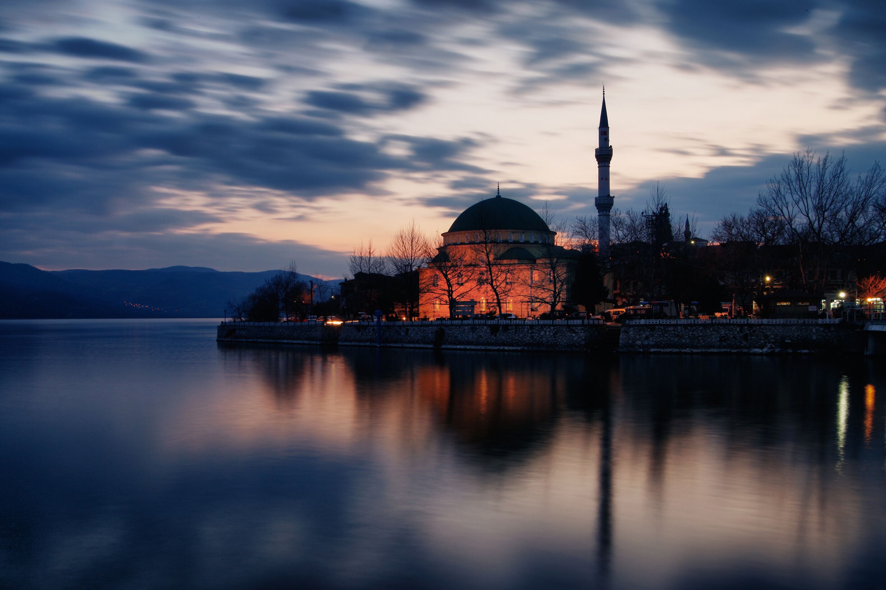 A mosque with a dome and minaret by a calm body of water at dusk.