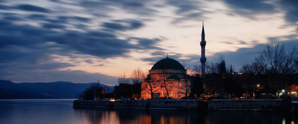 A mosque with a dome and minaret by a calm body of water at dusk.