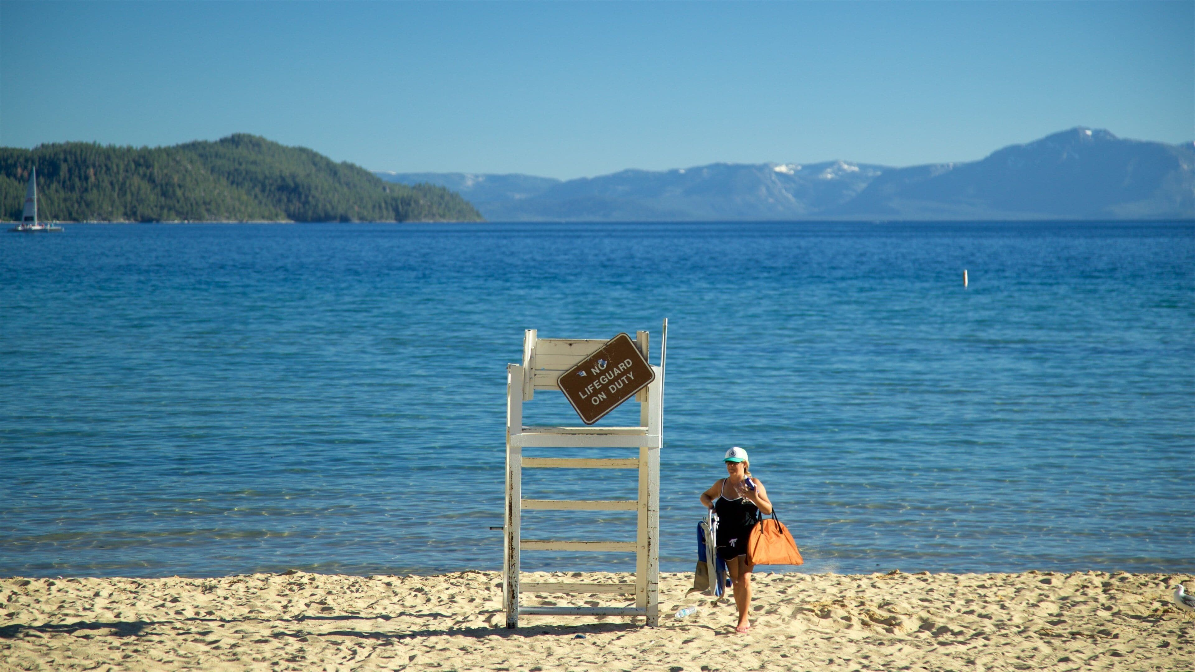Una playa con una silla de salvavidas y un cartel, una mujer caminando y un velero en el agua.