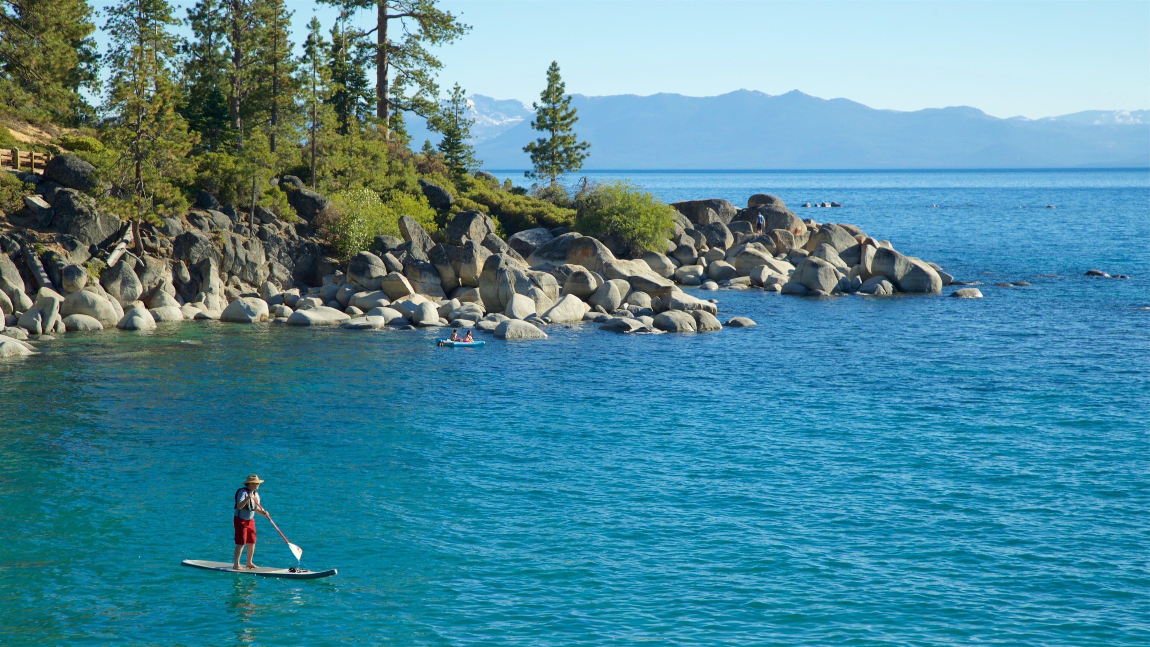 A person paddleboarding near a rocky shoreline with trees and mountains in the background.