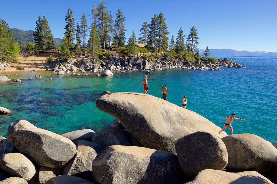 A rocky shoreline with clear blue water, people standing on rocks, and a forested hill in the background.