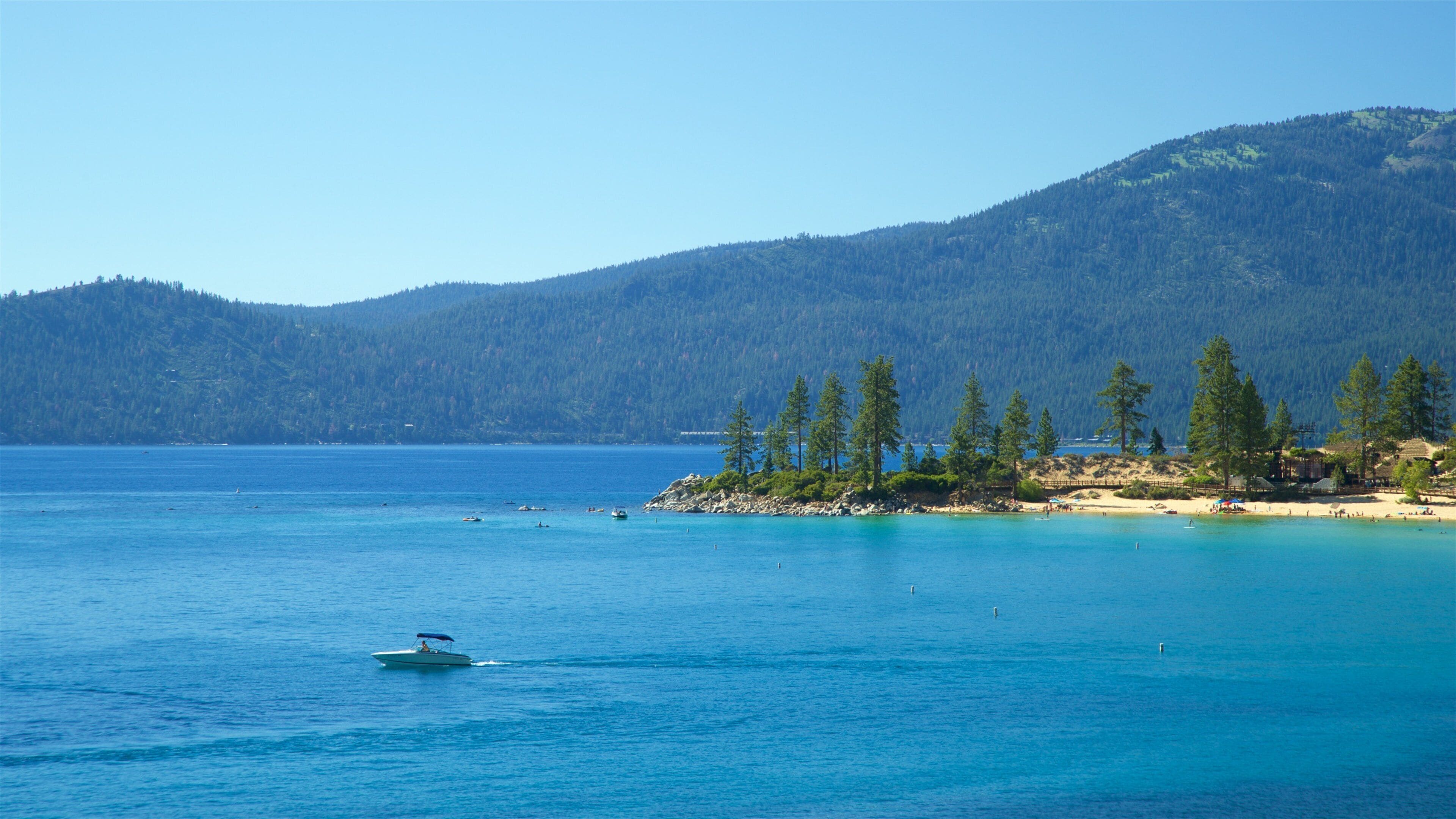 A clear blue lake with a boat, a sandy beach, and a forested mountain in the background.