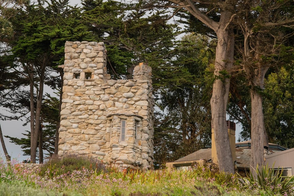 Una torre de piedra con un pequeño ventanal, rodeada de árboles y plantas florecientes.
