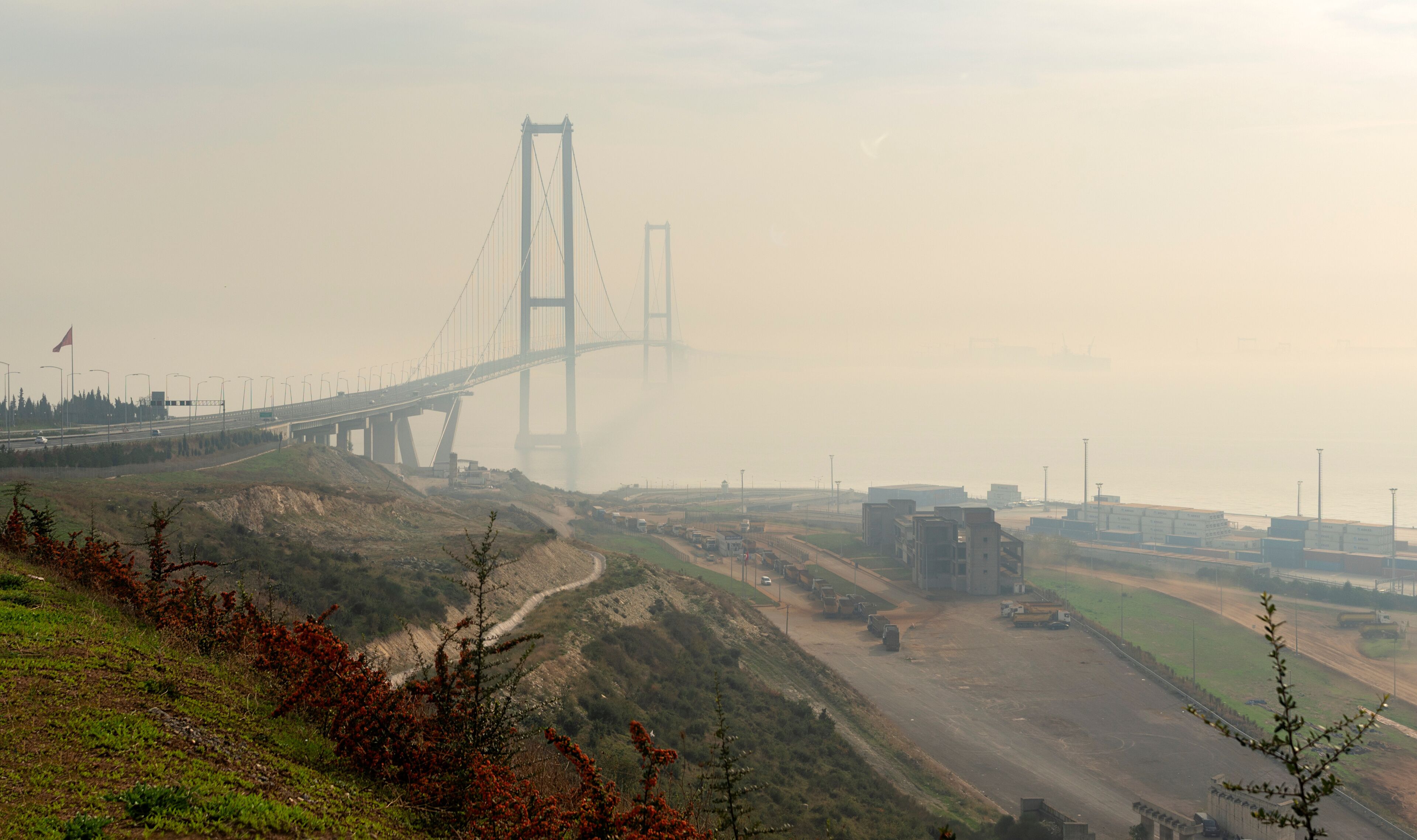 A hazy view of a large suspension bridge with multiple lanes, surrounded by a hilly landscape with sparse vegetation and industrial buildings in the background.
