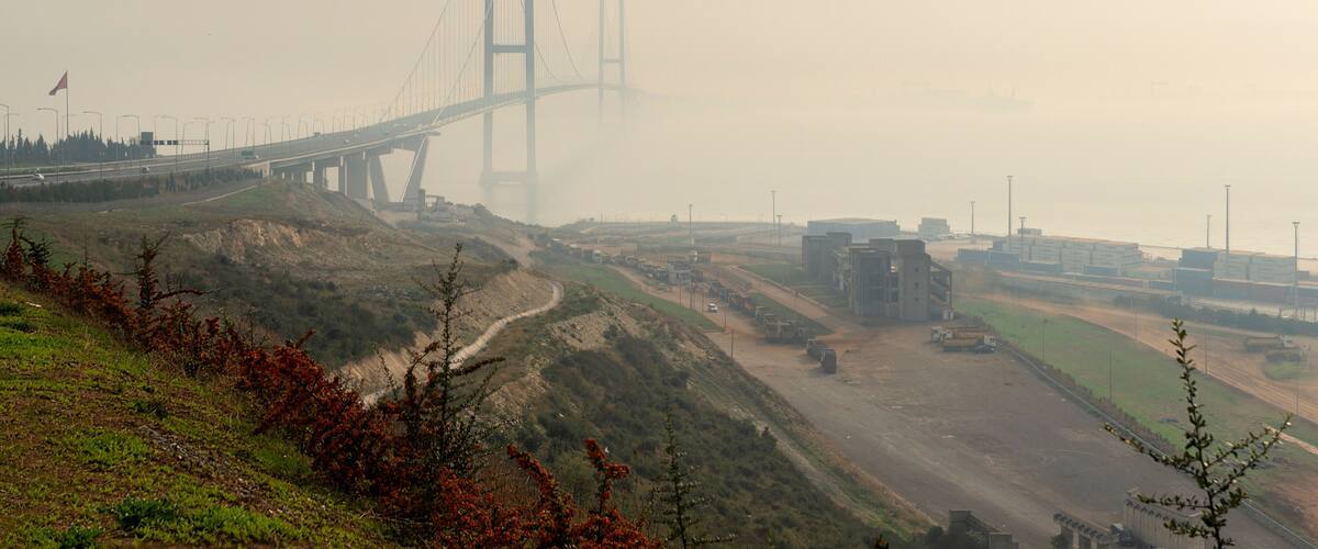 A hazy view of a large suspension bridge with multiple lanes, surrounded by a hilly landscape with sparse vegetation and industrial buildings in the background.