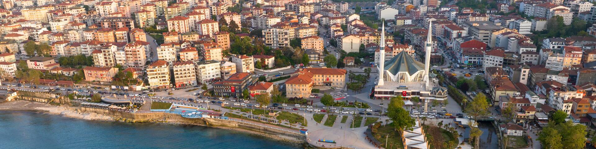 An aerial view of a coastal city with dense urban buildings, a prominent white church, and a circular stone structure in the water.