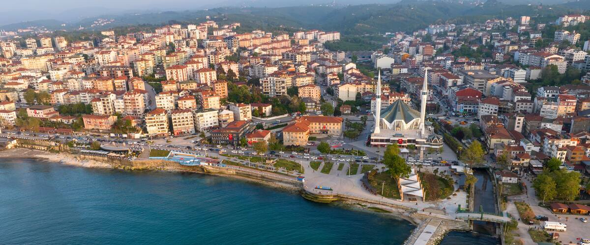 An aerial view of a coastal city with dense urban buildings, a prominent white church, and a circular stone structure in the water.