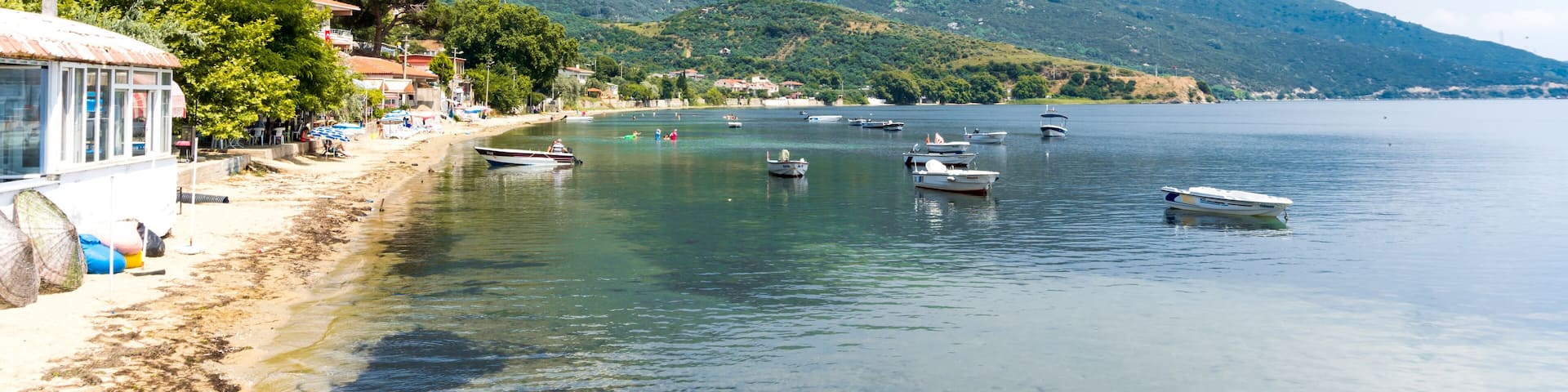A coastal scene with boats on calm water, a sandy beach, and a mountainous backdrop.