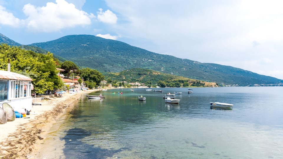 A coastal scene with boats on calm water, a sandy beach, and a mountainous backdrop.