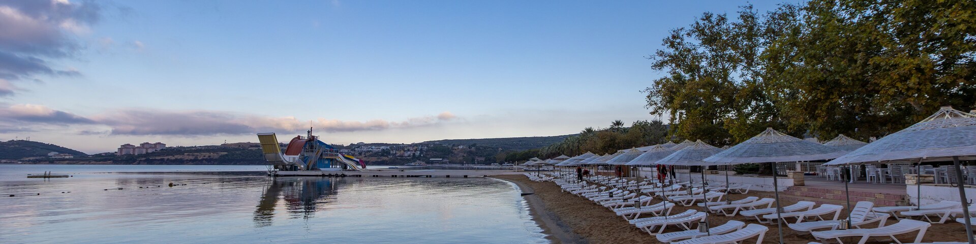 Una playa tranquila con sillas de descanso y sombrillas, un tobogán acuático y un horizonte urbano distante.