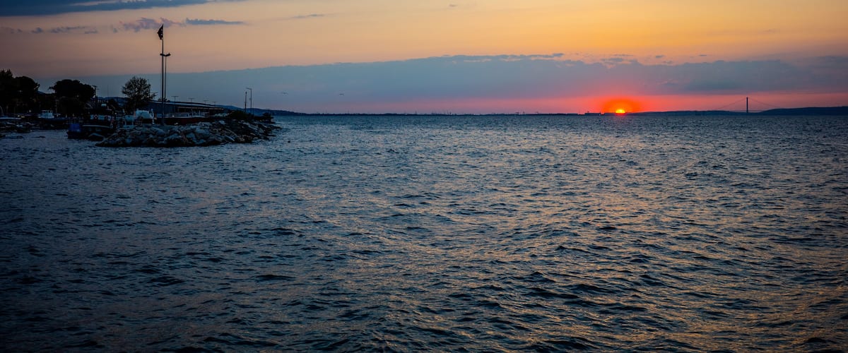A sunset over a calm sea with a pier and a bridge in the distance.