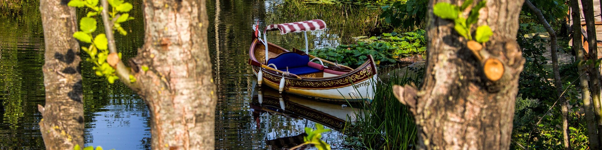 A traditional boat with a striped canopy docked in a calm river surrounded by trees and greenery.