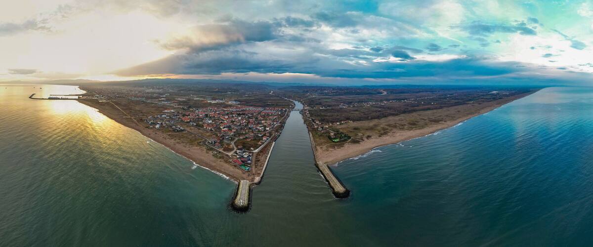 An aerial view of a coastal town with a prominent breakwater, a river running through the center, and a distant mountain range.