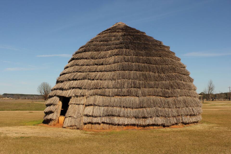 Une petite cabane en forme de dôme, recouverte de chaume, se dresse au milieu d’un champ d’herbe.