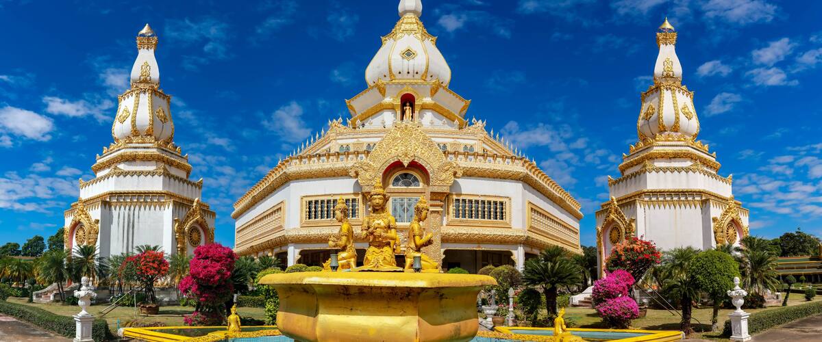 A large white and gold temple with a central statue and two smaller spires, surrounded by a garden with pink flowers and a fountain.