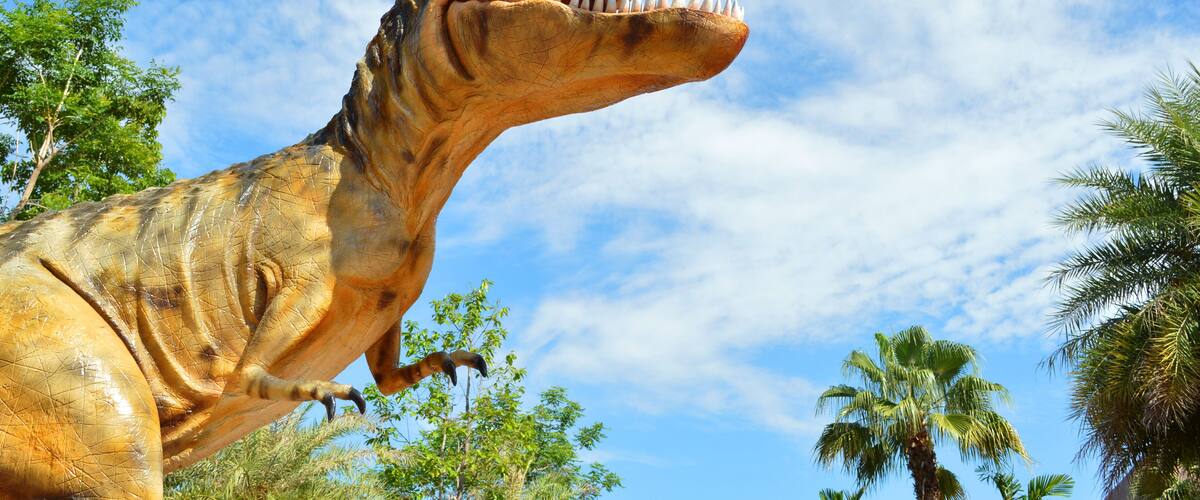 A large dinosaur statue with a wide-open mouth, set against a clear blue sky with scattered clouds.