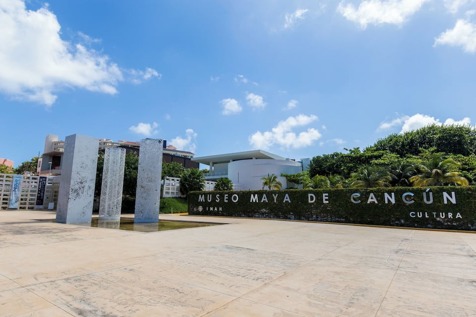 The Museo Maya de Cancún sign with a clear sky and palm trees in the background.