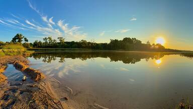 A calm lake with a clear reflection of the sky and trees, surrounded by grassy banks and a few scattered rocks.