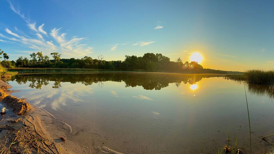 A calm lake with a clear reflection of the sky and trees, surrounded by grassy banks and a few scattered rocks.