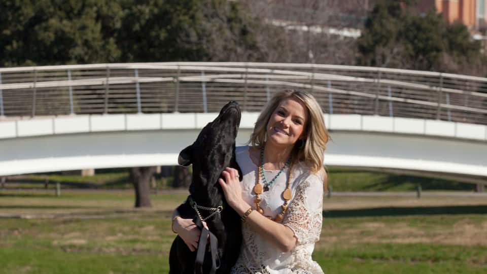 A person kneeling on grass with a dog, a white bridge, and tall buildings in the background.