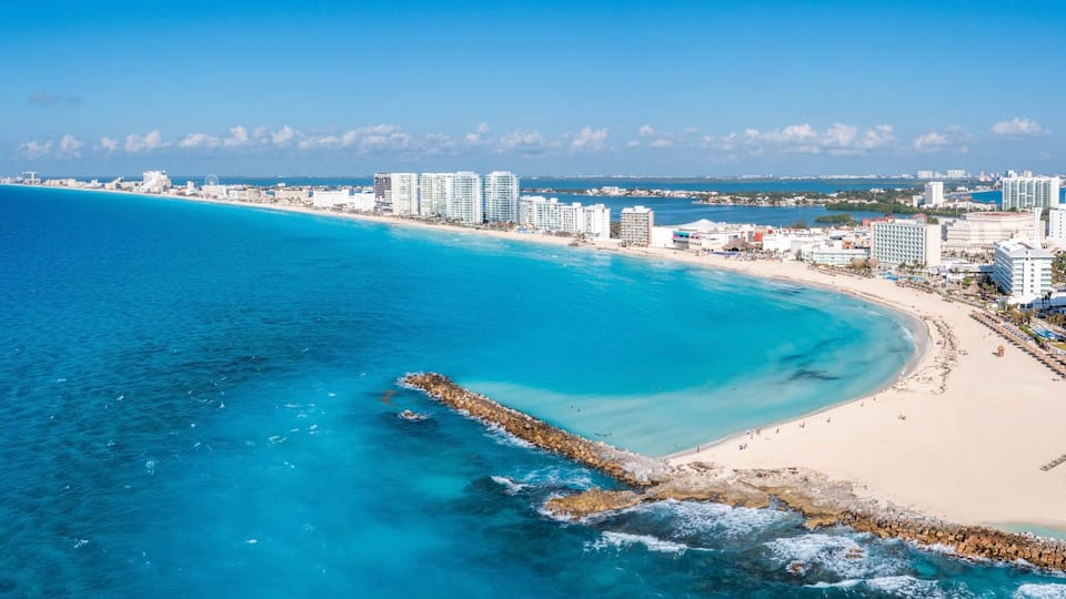 A coastal cityscape with a sandy beach, a rocky breakwater, and high-rise buildings along the shore.