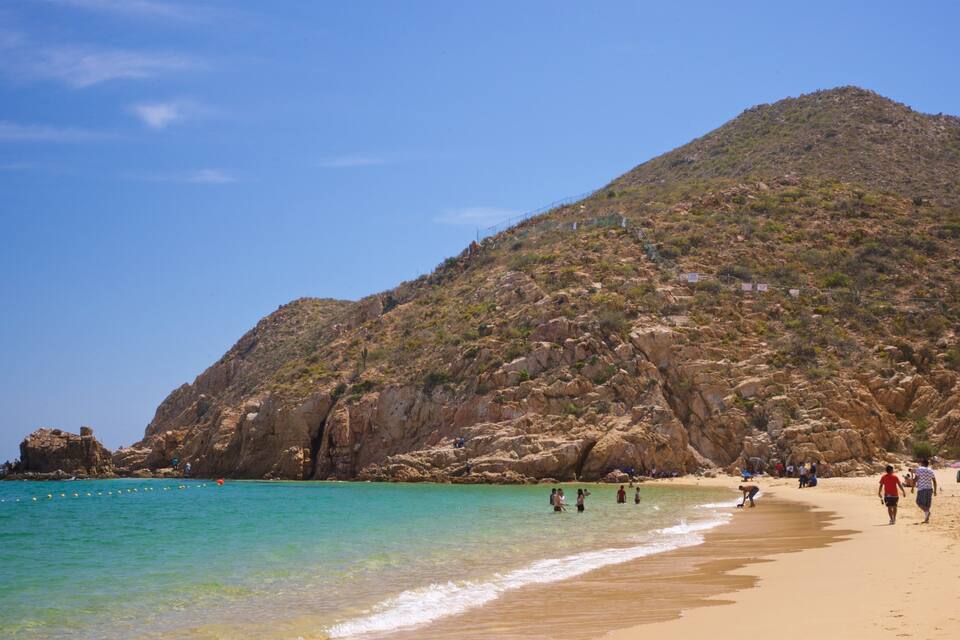 A beach with clear turquoise water, a sandy shore, and a rocky hill in the background.