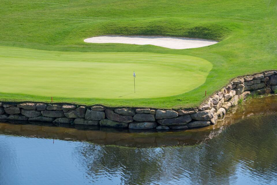 A golf course with a sand bunker, a flagstick, and a stone wall beside a body of water.