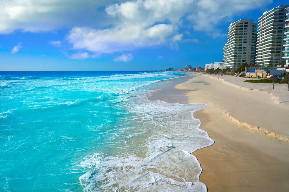 A beach with turquoise waves, sandy shore, and tall buildings in the background.