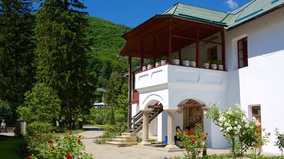 A white building with a wooden balcony, surrounded by lush greenery and flowering plants.