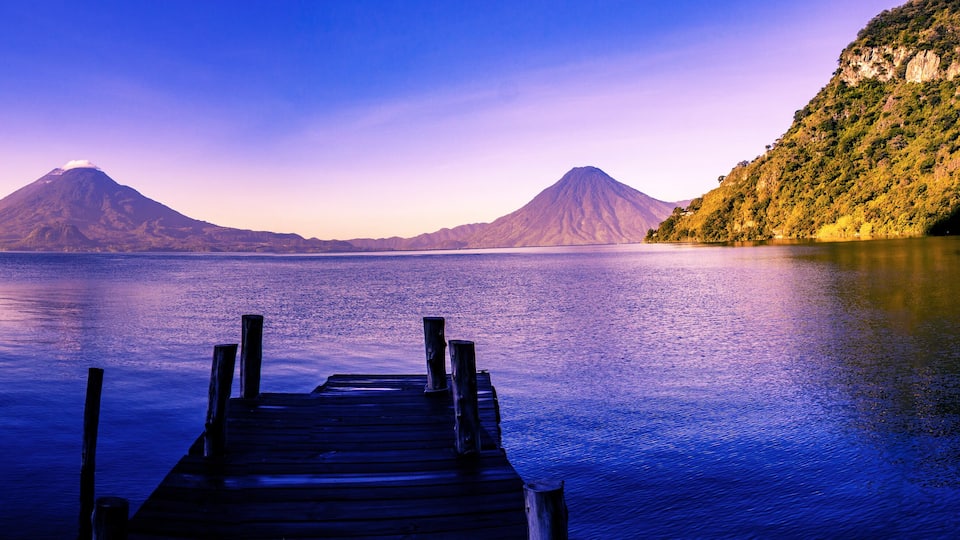 A wooden dock extending into calm waters with two prominent mountains in the background.