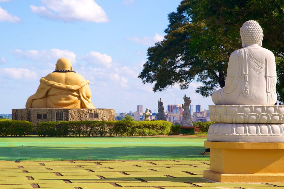 Um parque com duas grandes estátuas de Buda, uma sentada e outra em pé, rodeado por vegetação e um horizonte urbano ao fundo.