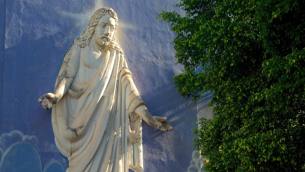 A statue of a robed figure with outstretched arms, set against a blue wall with cloud patterns.