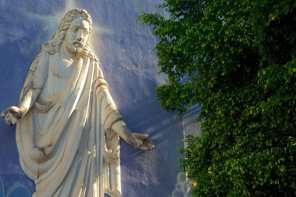Een standbeeld van een gewaande figuur met uitgestrekte armen, voor een blauwe muur met wolkenpatronen.