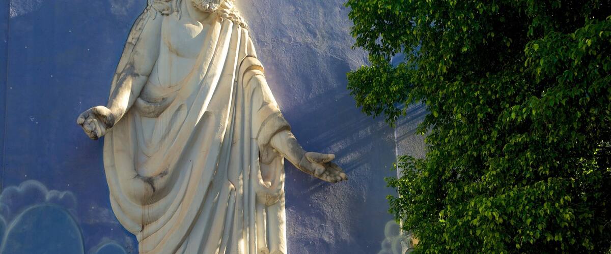 A statue of a robed figure with outstretched arms, set against a blue wall with cloud patterns.