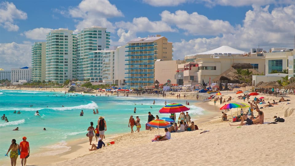 A crowded beach with people swimming and relaxing under colorful umbrellas, surrounded by tall hotels and buildings.