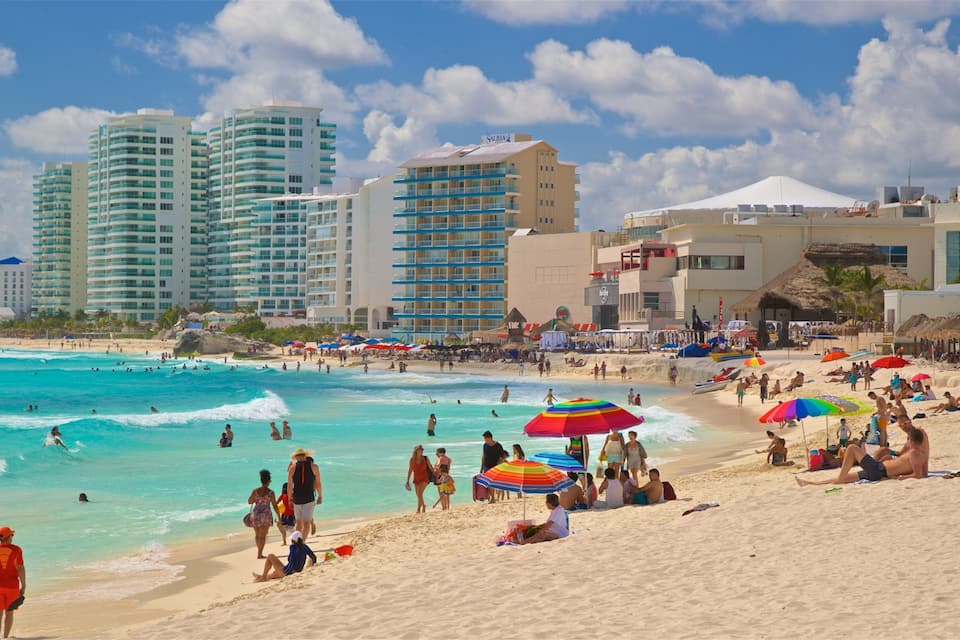 Une plage bondée où les gens se baignent et se détendent sous des parasols colorés, entourée de grands hôtels et d’immeubles.