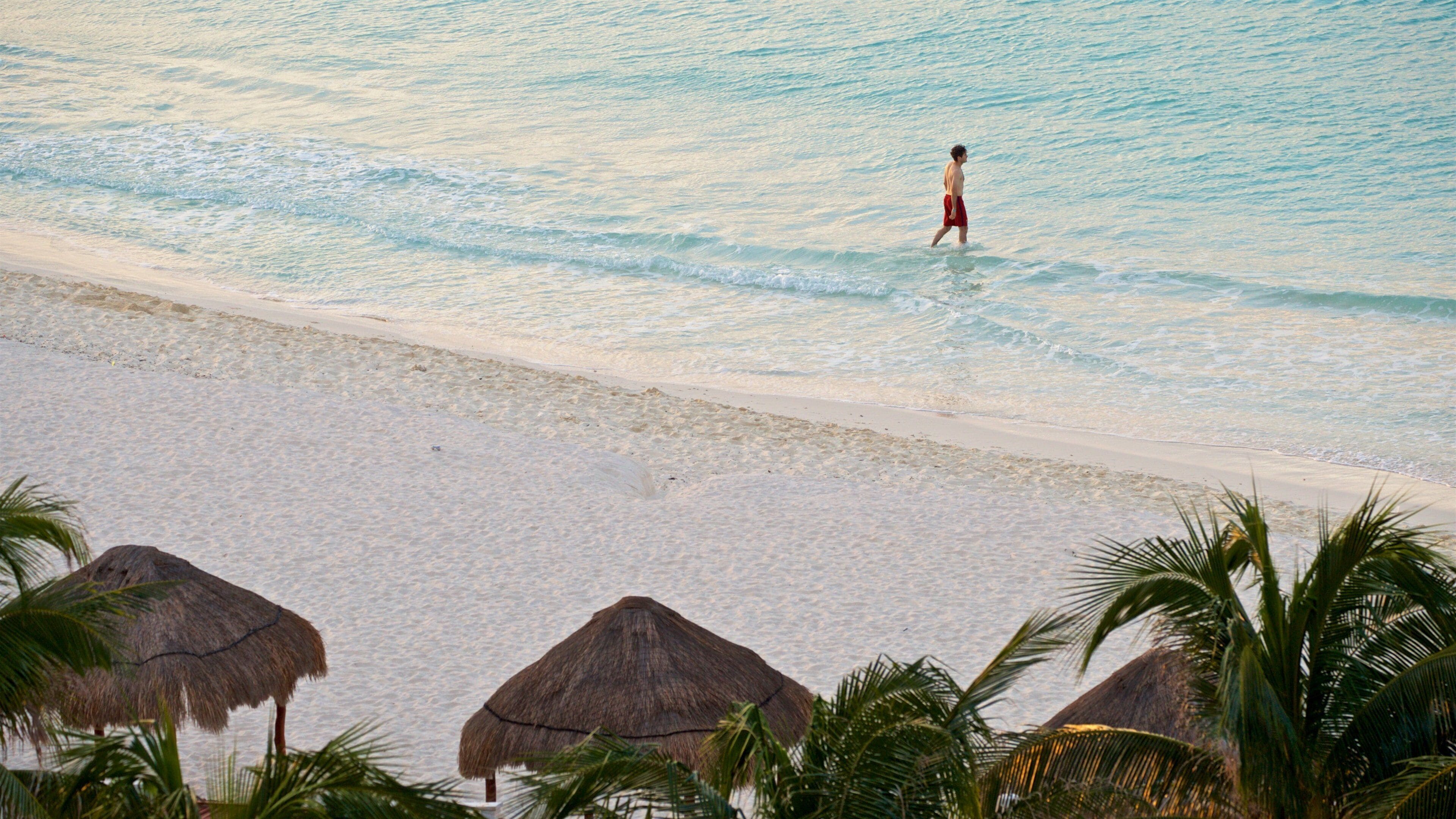 A beach with clear water, a person walking, and thatched huts.