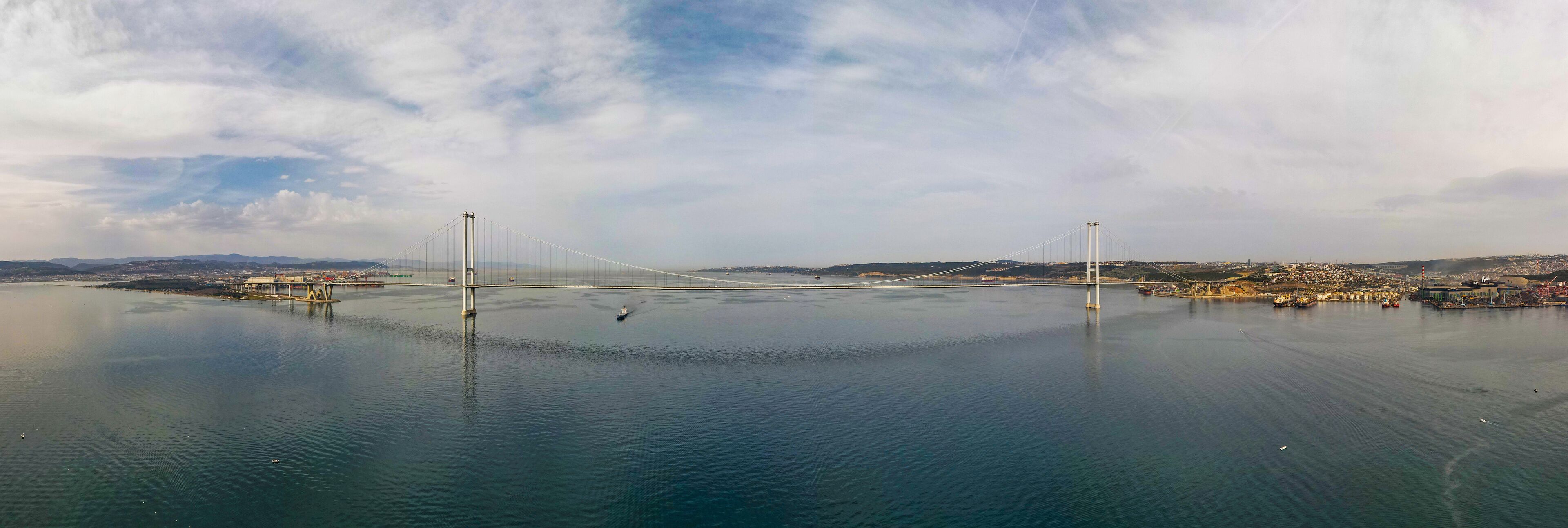 A large suspension bridge spanning a calm body of water with a cityscape in the background.